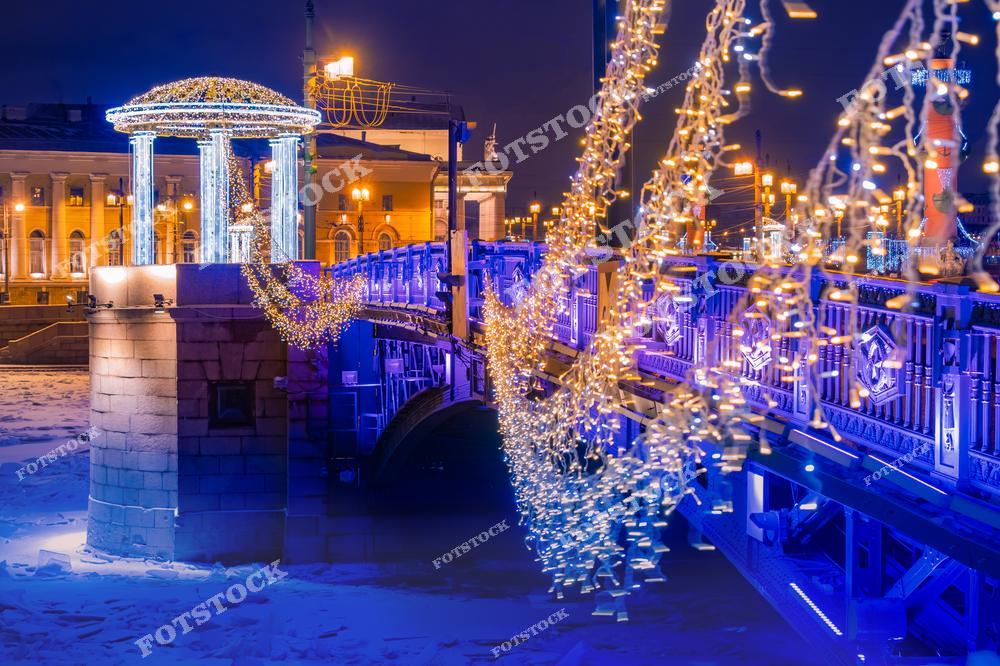 На фотографии изображен Дворцовый мост в Санкт-Петербурге, украшенный новогодними гирляндами и подсветкой. Колонны на мосту освещены яркими огнями, подчеркивающими их элегантность. На заднем плане виднеются Ростральные колонны и здания на Стрелке Васильевского острова. Ледяная гладь Невы и ночное освещение создают праздничное настроение.